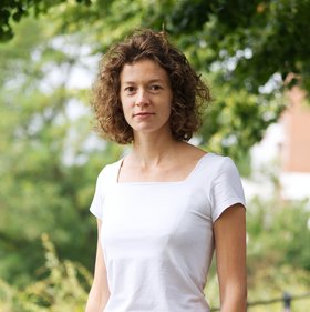 Dr. Nathalie Vielle: eine lächelnde Forscherin mit lockigen Haaren in einem weißen T-Shirt auf einem sonnigen Weg unter Bäumen