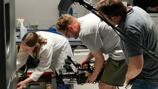 A researcher can be seen placing a sample in an incubator. Behind her is a cameraman with a camera and a presenter. A microphone can also be seen.