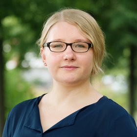Dr Lisa Oestereich: a researcher with brown glasses, blond hair tied in a braid and blue blouse, green branches in the background.