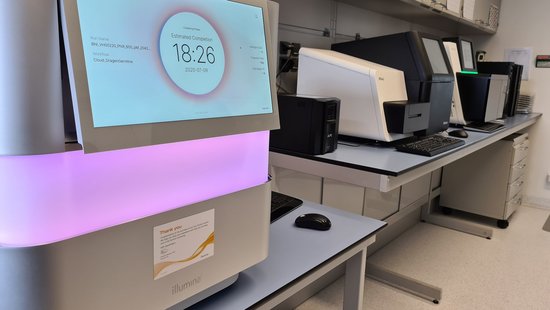 The Next-Generation Sequencing (NSG) unit of the BNITM can be seen. In the foreground is a large white box on a table. In the middle runs a wide luminous strip that glows pink-purple. At the top, you can see a large screen with the text estimated completion 18:26. In the background, there is more laboratory equipment on tables.