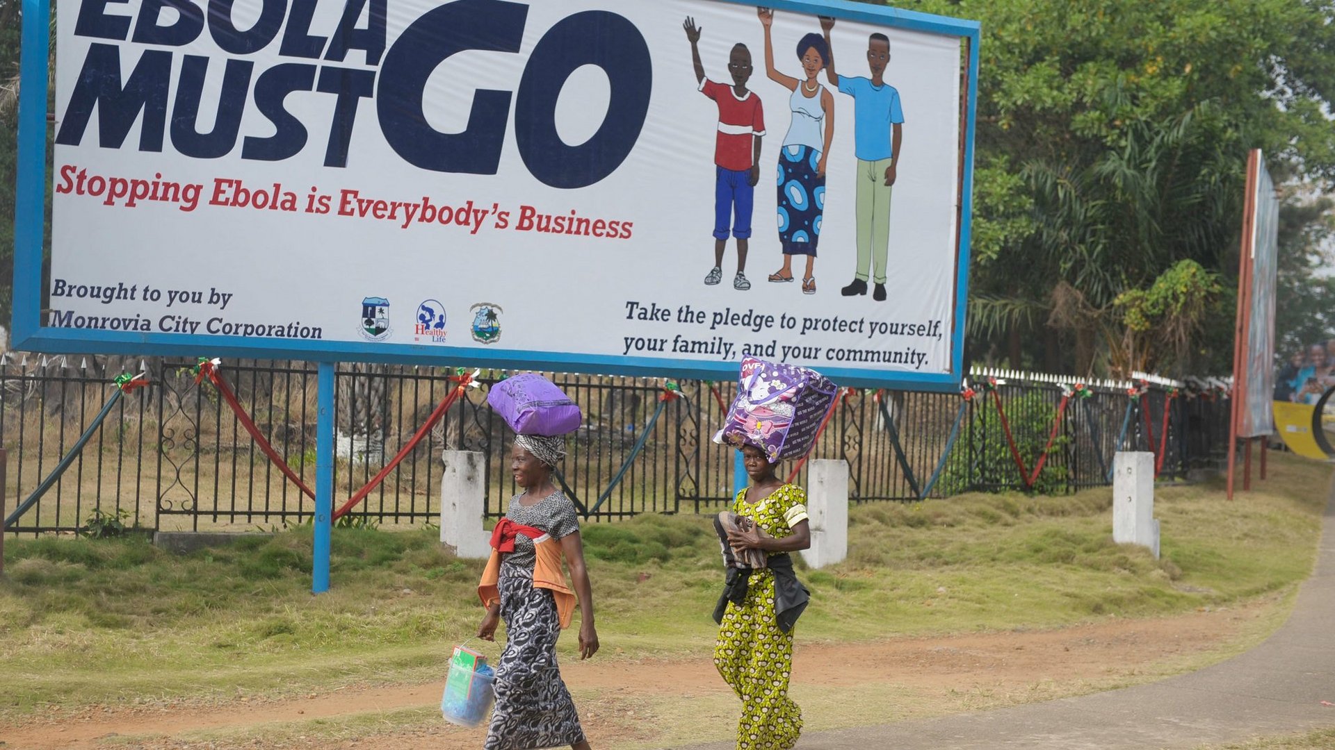 The picture shows two women walking in the streets of Monrovia, the capital of Liberia. They wear colorful scarves and have bags on their heads. In the background is a large street poster encouraging citizens of Monrovia to help fight Ebola.