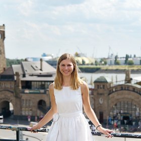 Dr Kerstin Unfried: a researcher with shoulder-length blonde hair and a white dress stands in front of the ancient building of the Landungsbrücken.