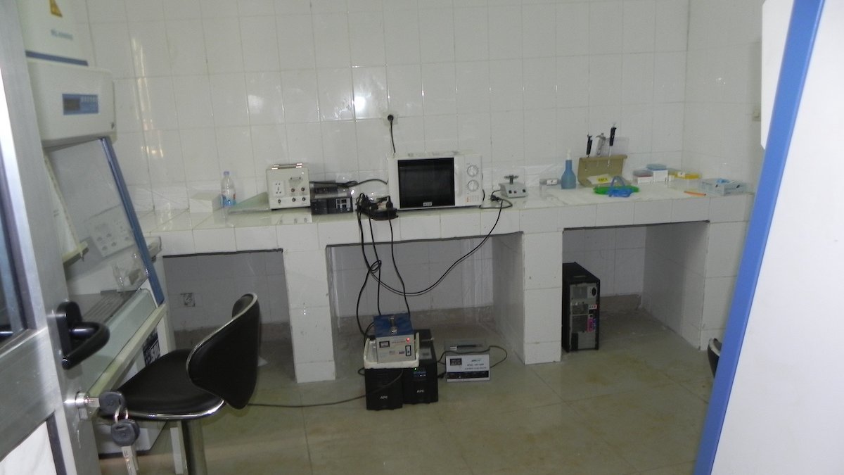 Das Bild zeigt ein Labor. Link steht eine Sterilbank, an der Wand geradeaus eine TIsch mit Computer, Mikrowelle und anderen unterschiedlichen Laborgegenständen.
