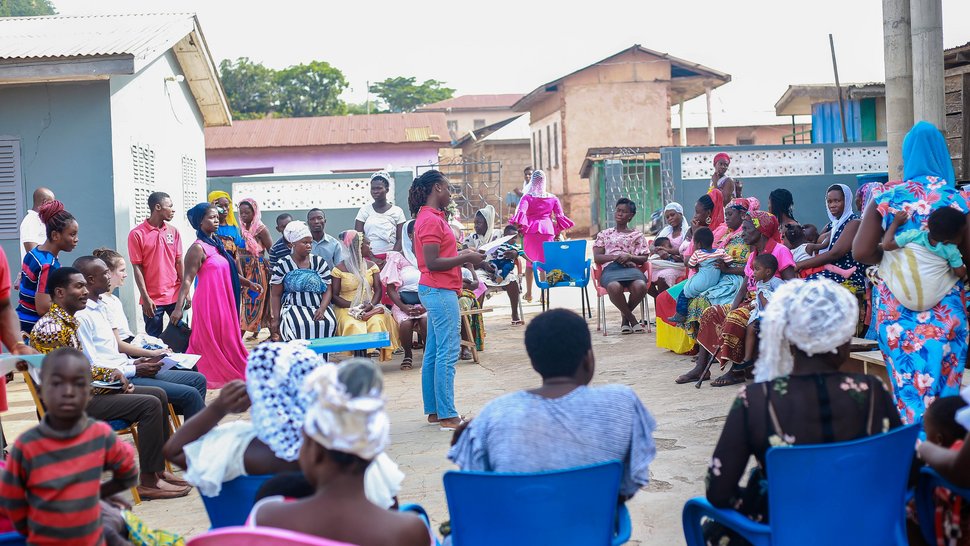 The photo shows a village meeting outside. A health worker is standing in the centre explaining something.