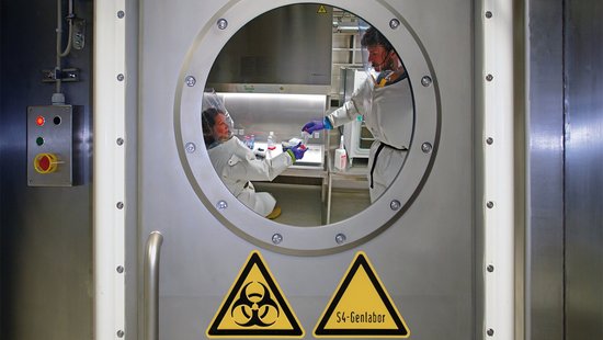 You can see a metal end door with two yellow triangular warning signs on it. One shows the biohazard symbol, the second says S4 genetic laboratory. You can look inside through a round window and see two researchers inside. Both are wearing a full white suit and a transparent cap on their heads. One researcher is sitting at a workbench while the second researcher hands her a transparent cell culture flask containing red cell medium.  Both researchers are wearing a headset.