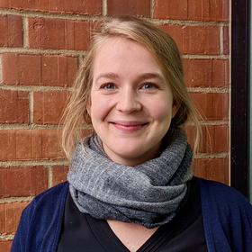 PhD Silke Olschewski-Pavlita: A researcher smiles into the camera. She wears her blond hair loose in a plait with a side parting, a grey scarf and a black shirt with a blue cardigan.