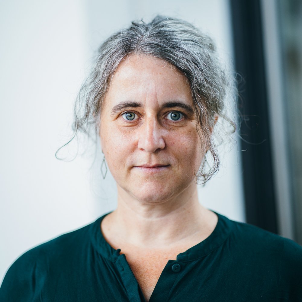 Portrait of a white female person with brown-grey hair pulled back, wearing a dark green shirt
