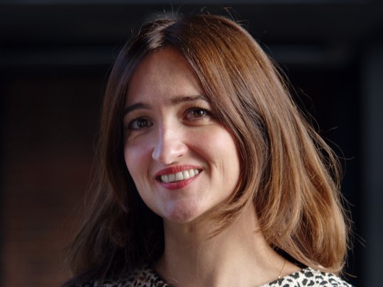 Dr Denise Dekker: a researcher with long brown hair, wearing a black and white patterned blouse. She stands in front of a dark background.