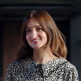 Dr Denise Dekker: a researcher with long brown hair wearing a black and white patterned blouse. She stands in front of a dark background.