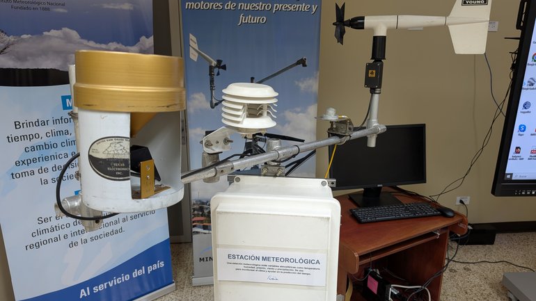 A weather station display at Costa Rica's National Meteorological Institute (IMN), featuring a rain gauge, sensor array, wind vane, and data logger.