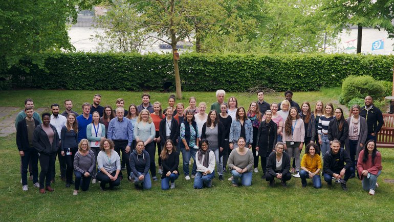 [Translate to English:] The picture shows about 50 people of different ages and looks standing or squatting on a meadow. Behind them is a horizontal line: a row of bushes, behind which the Elbe can be guessed.