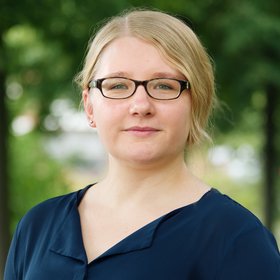 Dr. Lisa Oestereich: eine Forscherin mit brauner Brille, blonden Haaren, die zu einem Zopf gebunden sind und blauer Bluse, im Hintergrund grüne Äste.