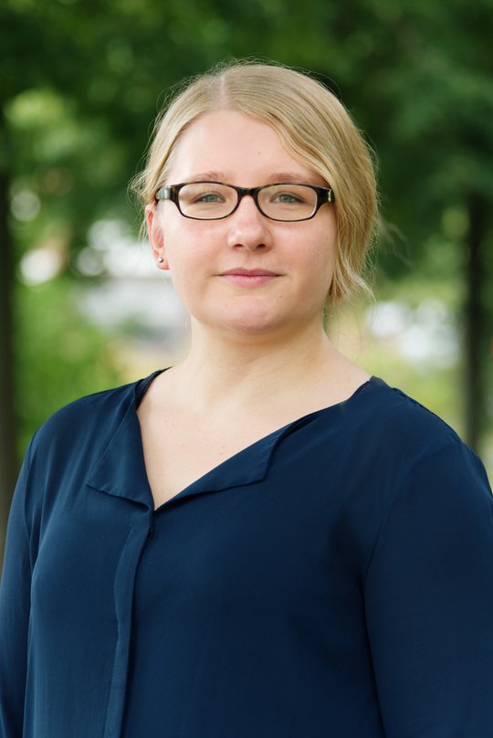 Dr. Lisa Oestereich Dr. Lisa Oestereich: eine Forscherin mit brauner Brille, blonden Haaren, die zu einem Zopf gebunden sind und blauer Bluse, im Hintergrund grüne Äste.
