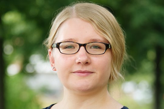 Dr Lisa Oestereich: a female researcher with brown glasses, blonde hair tied back, wearing a blue blouse, with green branches in the background.