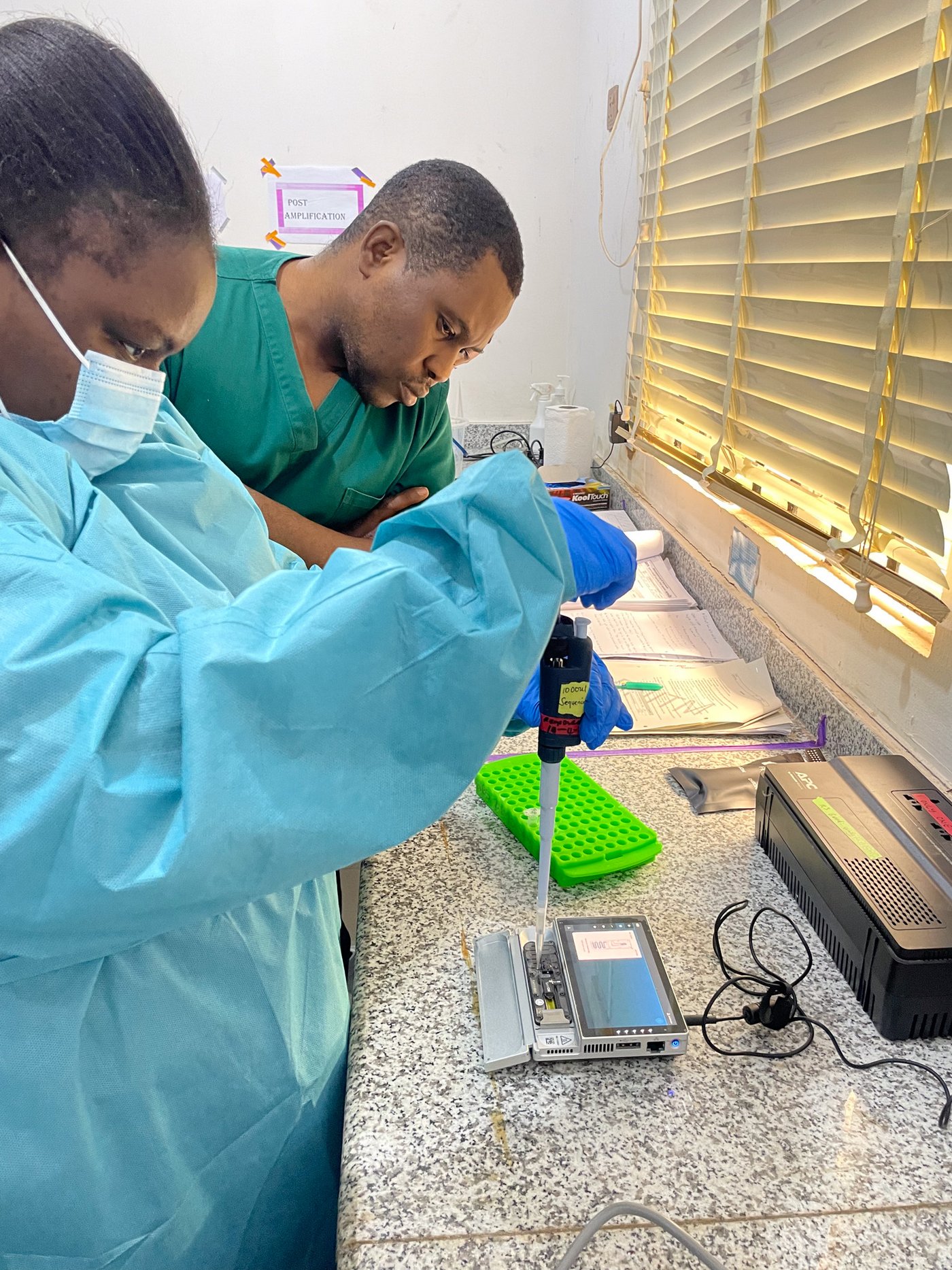 Das Foto zeigt zwei nigerianische Experten (weiblich und männlich) in blauen bzw. grünen Laboranzügen beim Laden von Proben für die metagenomische Sequenzierung auf der MinION Nanopore Plattform.