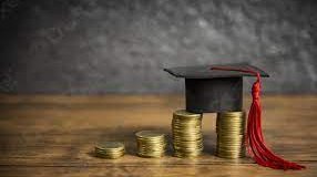 Graduation hat on money coins on a table in front of a dark background.
