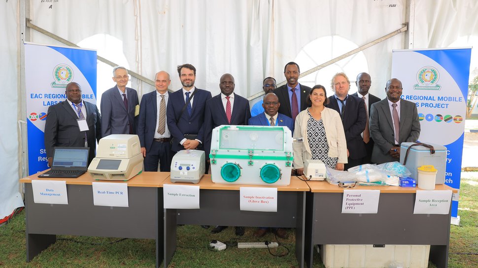Eröffnung UVRI Exzellenzzentrum Auf dem Foto ist ein Tisch zusehen, auf dem Geräte wie eine PCR-Maschine und eine Glove Box stehen. Dahinter stehen 12 gut gekleidete Personen. Flankiert werden sie von zwei Postern des EAC Mobile Lab Projektes.
