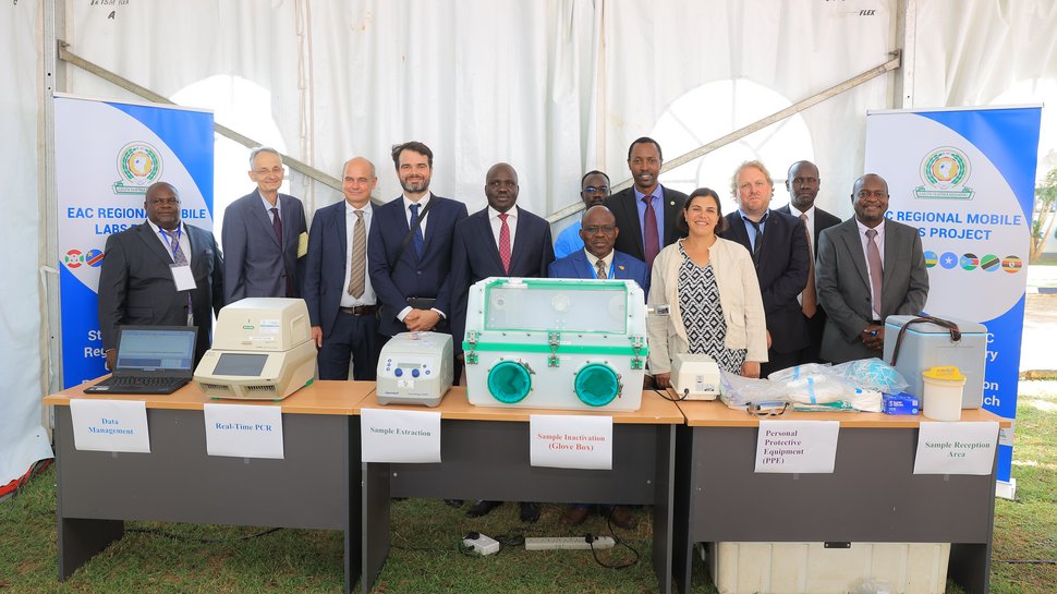 Auf dem Foto ist ein Tisch zusehen, auf dem Geräte wie eine PCR-Maschine und eine Glove Box stehen. Dahinter stehen 12 gut gekleidete Personen. Flankiert werden sie von zwei Postern des EAC Mobile Lab Projektes.