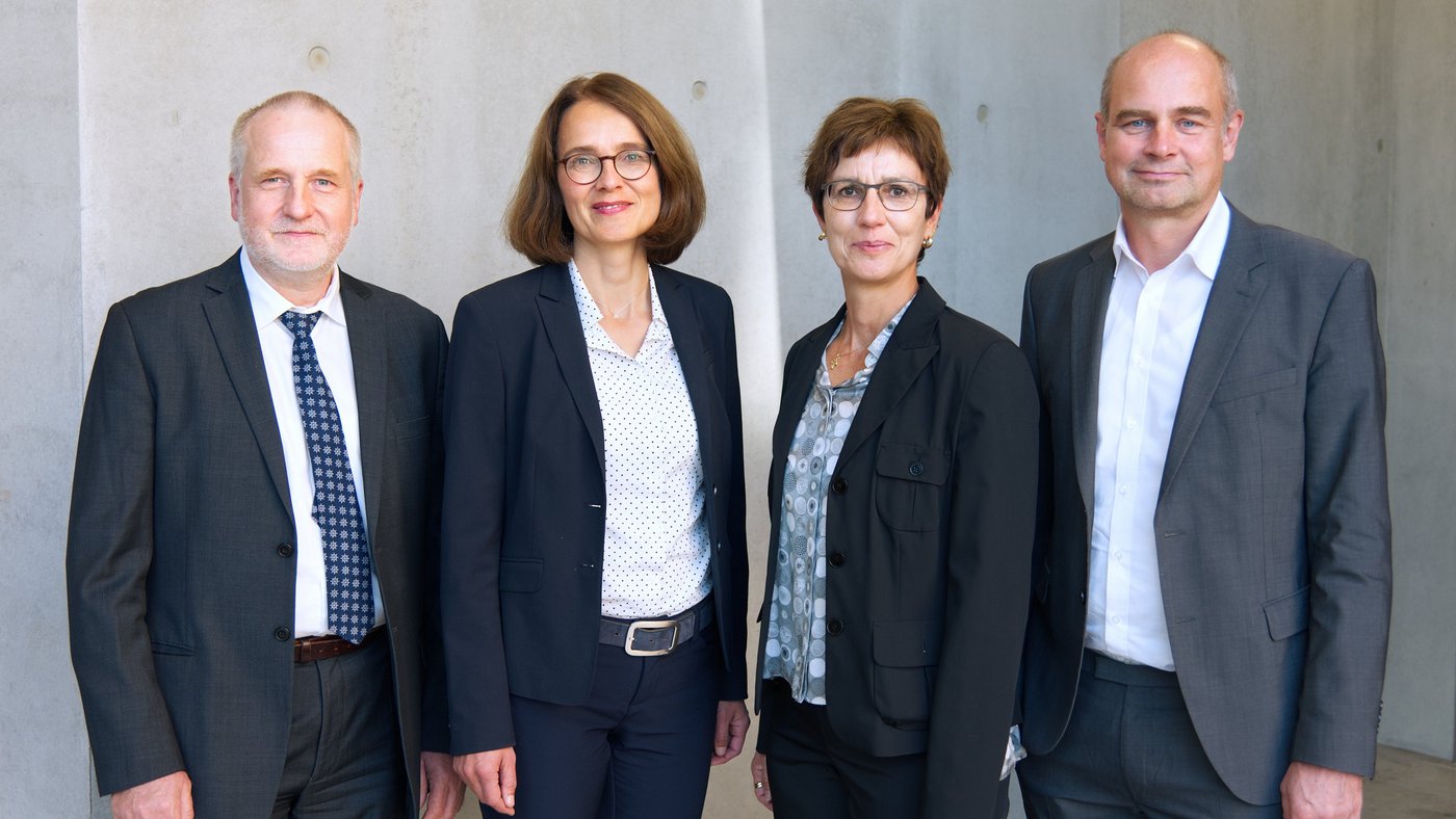 Foto der vier Vorstandsmitglieder: Zwei Frauen und zwei Männer stehen in dunklen Kostümen bzw. Anzügen gekleidet vor einer Betonwand und lächeln Richtung Kamera.