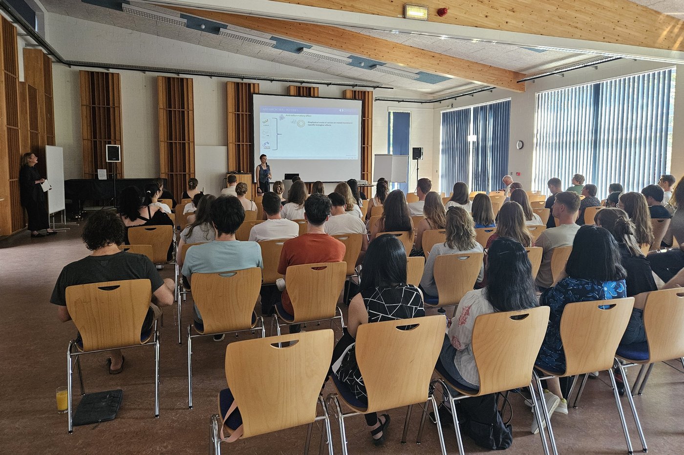 LCI Summer School Blick von hinten auf Studierenden, die einem Vortrag in einem hellen Seminarraum mit Holzdecke und großen Fenstern folgt. Zwei Personen stehen vorne und präsentieren auf einer Leinwand.