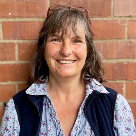 Carola Busch: A technical assistant looks into the camera. She has shoulder-length brown-grey hair with fringes. She has glasses tucked into her hair and is wearing a light blue floral blouse and a dark blue waistcoat.