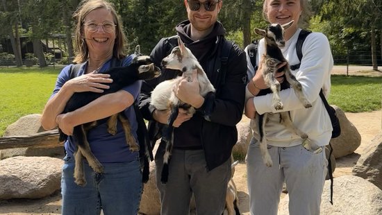 Wildpark Schwarzeberge Three people stand outdoors, each holding a baby goat, with trees and rocks in the background under a clear sky.
