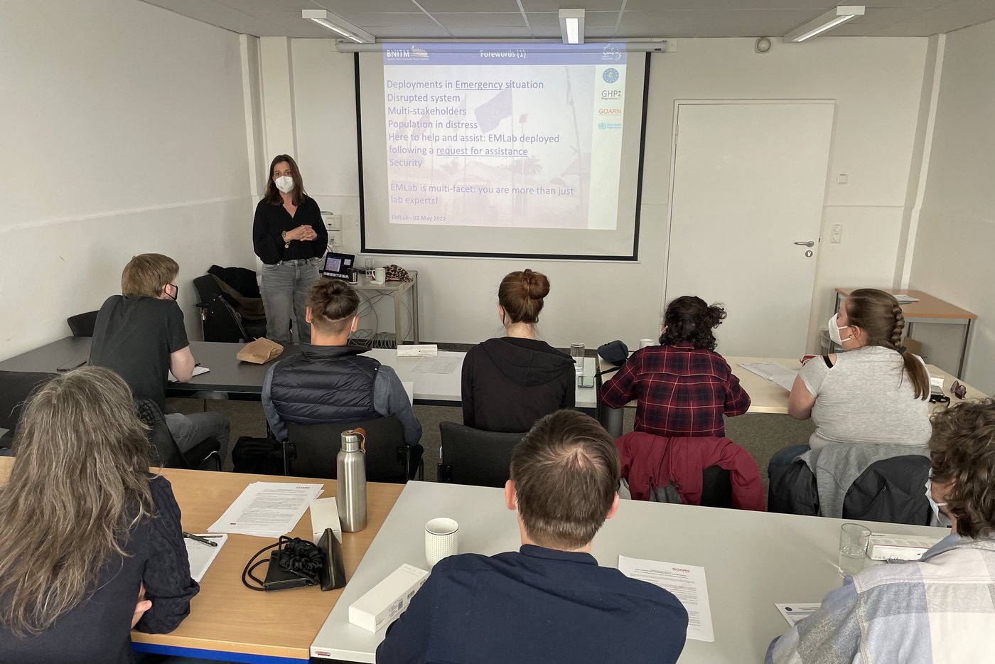 In the classroom: Several trainees listen to the lecturer standing in front of a screen.