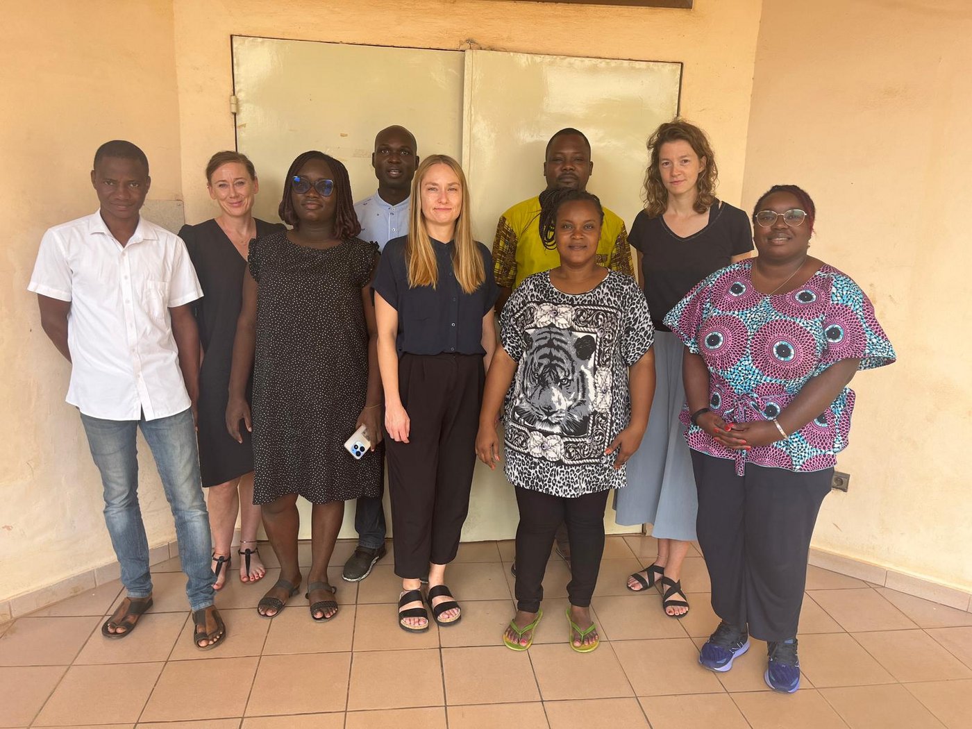The Parakou lab team with the BNITM team in Parakou, Oct 2023 A diverse group of nine people, partly in colourful African clothes, stands in front of an apricot-coloured wall with a metal door.