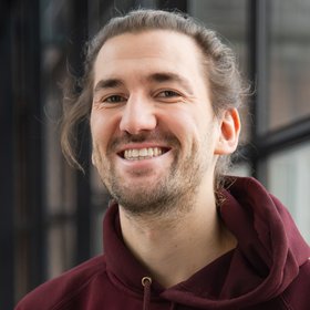 Nils Petersen Foto von Nils Petersen. Er hat hochgebundene, lange Haare und einen Bart, er trägt einen dunkelroten Kapuzenpullover und steht vor einer Fensterfront.