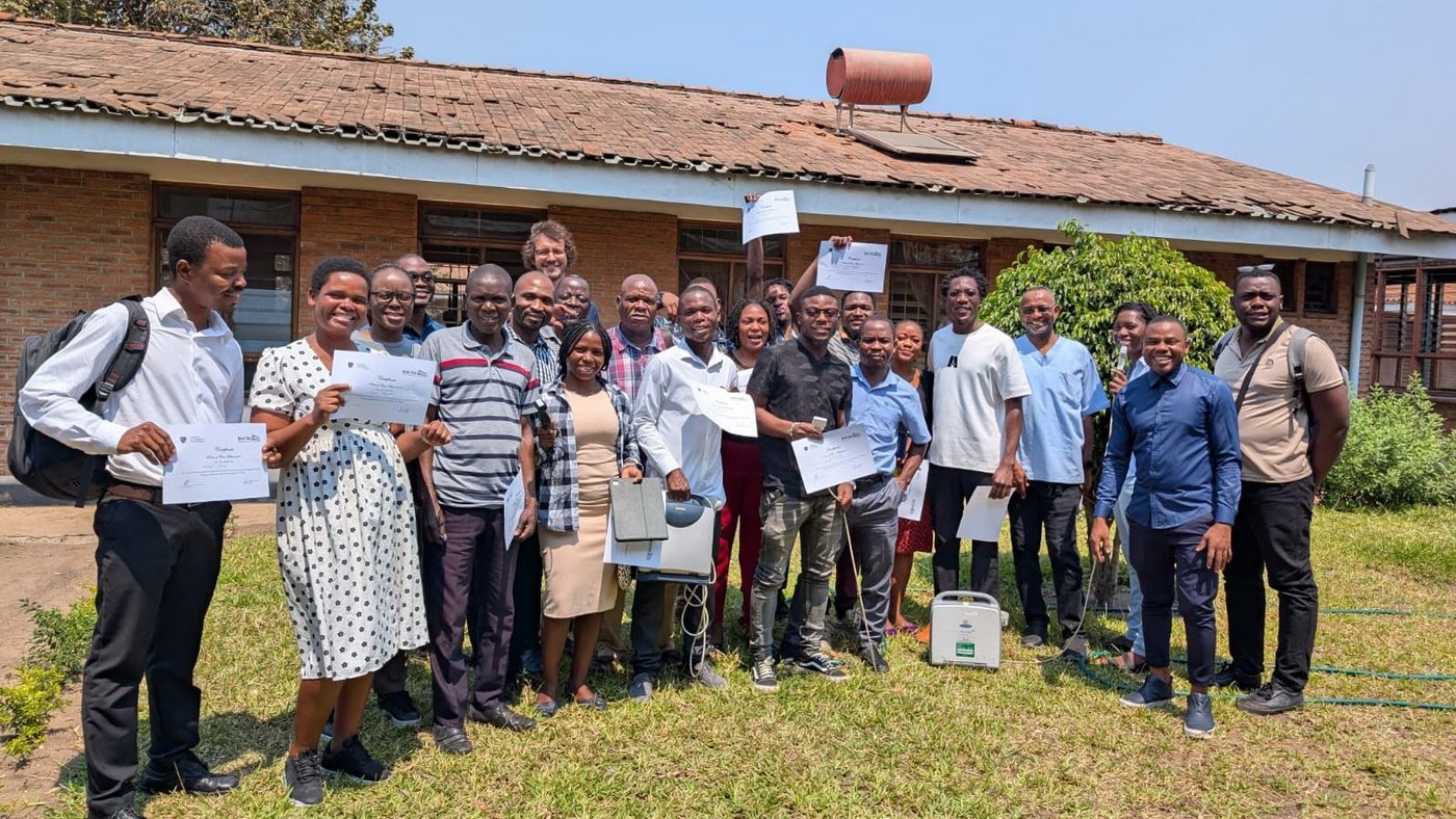 The picture shows participants and lecturers from the course in Mangochi standing in front of a building. The participants are holding their certificates.