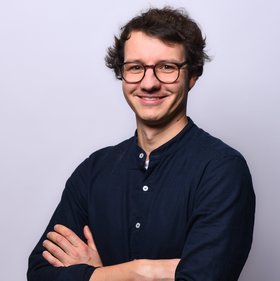 Das Bild zeigt einen braunhaarigen Mann, der ein dunkeles Hemd trägt, seine Arme verschränkt hat und freundlich in die Kamera schaut.