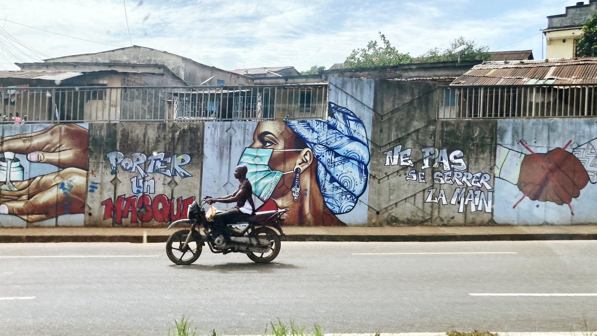 A male Beninese rides a motorbike. Graffiti on the wall behind him says “wear a mask” and “Do not shake hands” in French. Next to that you see illustrations of hands using sanitizer, a female Beninese wearing a mask and a handshake with a red cross over it.