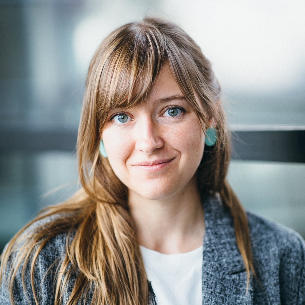 Portrait of a white female person with long brown hair, wearing a grey suit and a white shirt.