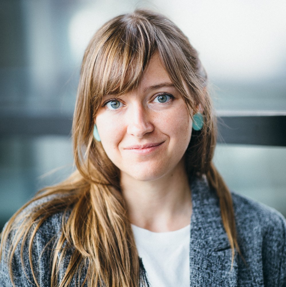 Portrait of a white female person with long brown hair, wearing a grey suit and a white shirt.