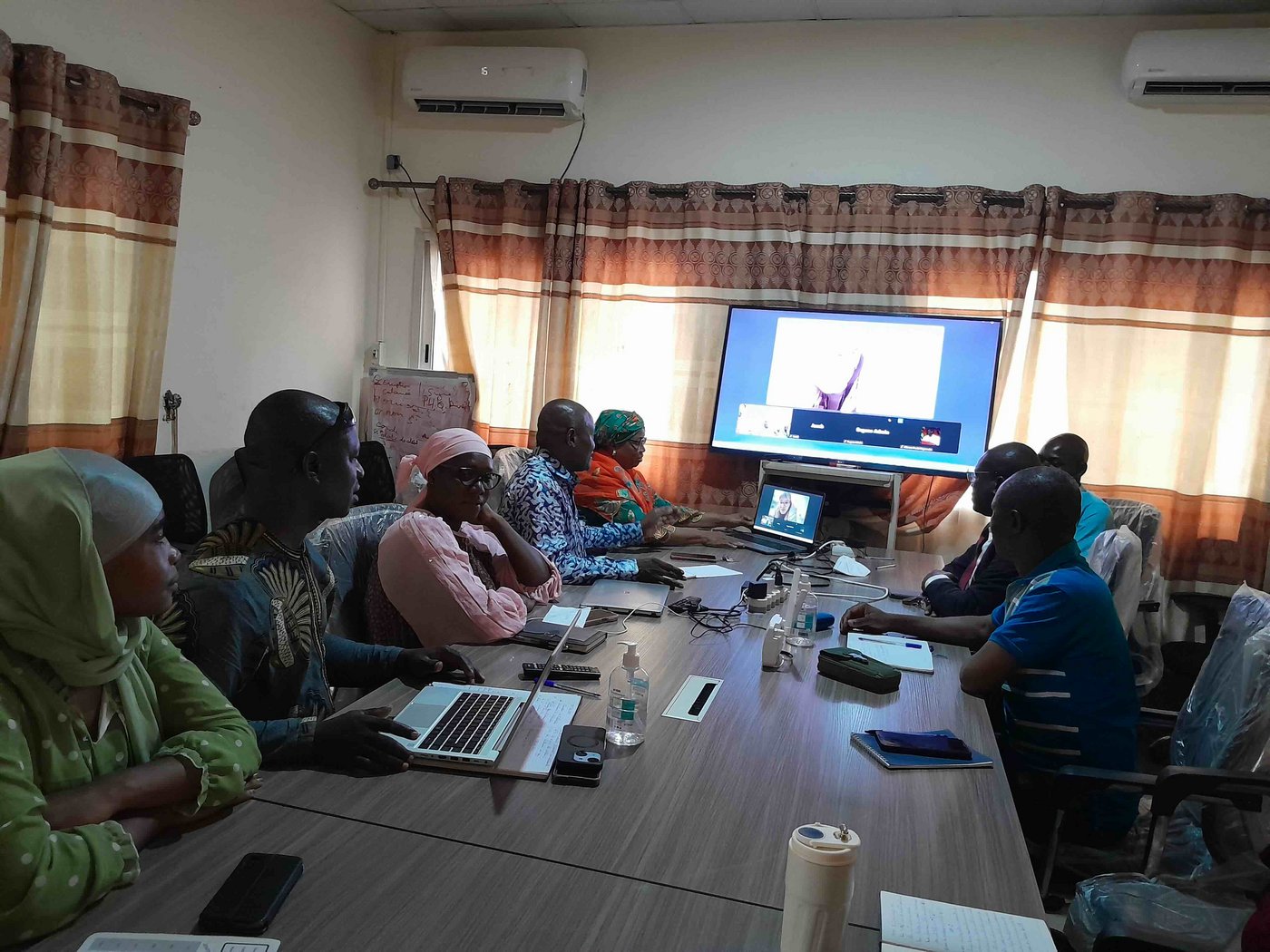 Remote training on yellow virus fever at CRV-LFHVG. Conakry, Guinea, August 2025 Several laboratory staff attending the remote training in the conference room. They are all seated around a big table and listening to an online presentation on a big screen.