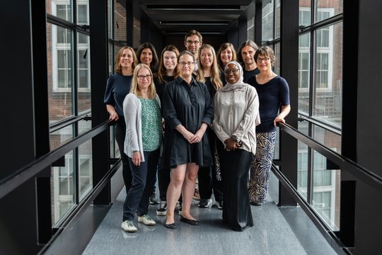 A diverse group of 11 people smiling in a glass-walled corridor. They stand in two rows, expressing teamwork and positivity.