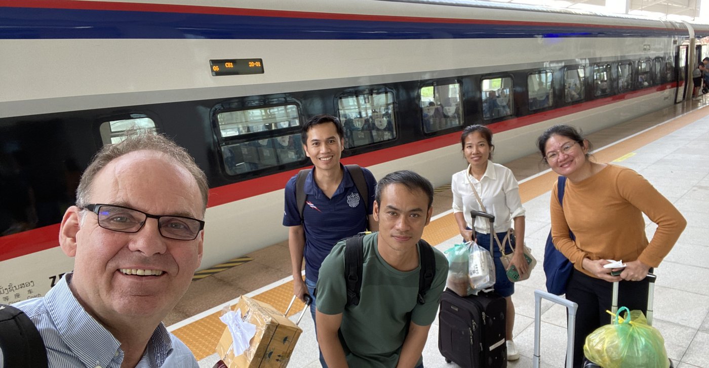 The picture shows five people standing in front of a train with luggage and looking friendly into the camera.