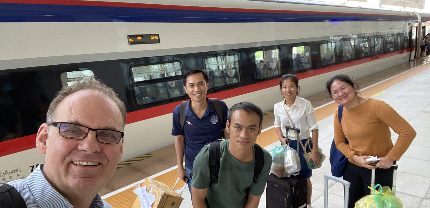 Das Bild zeigt fünf Menschen, die mit Gepäck vor einem Zug stehen und freundlich in die Kamera schauen.