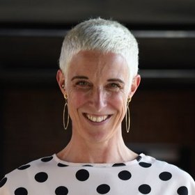 Dr Wiebke Hartmann: A researcher laughs into the camera, she wears short blonde hair, a white shirt with black dots and large gold earrings.