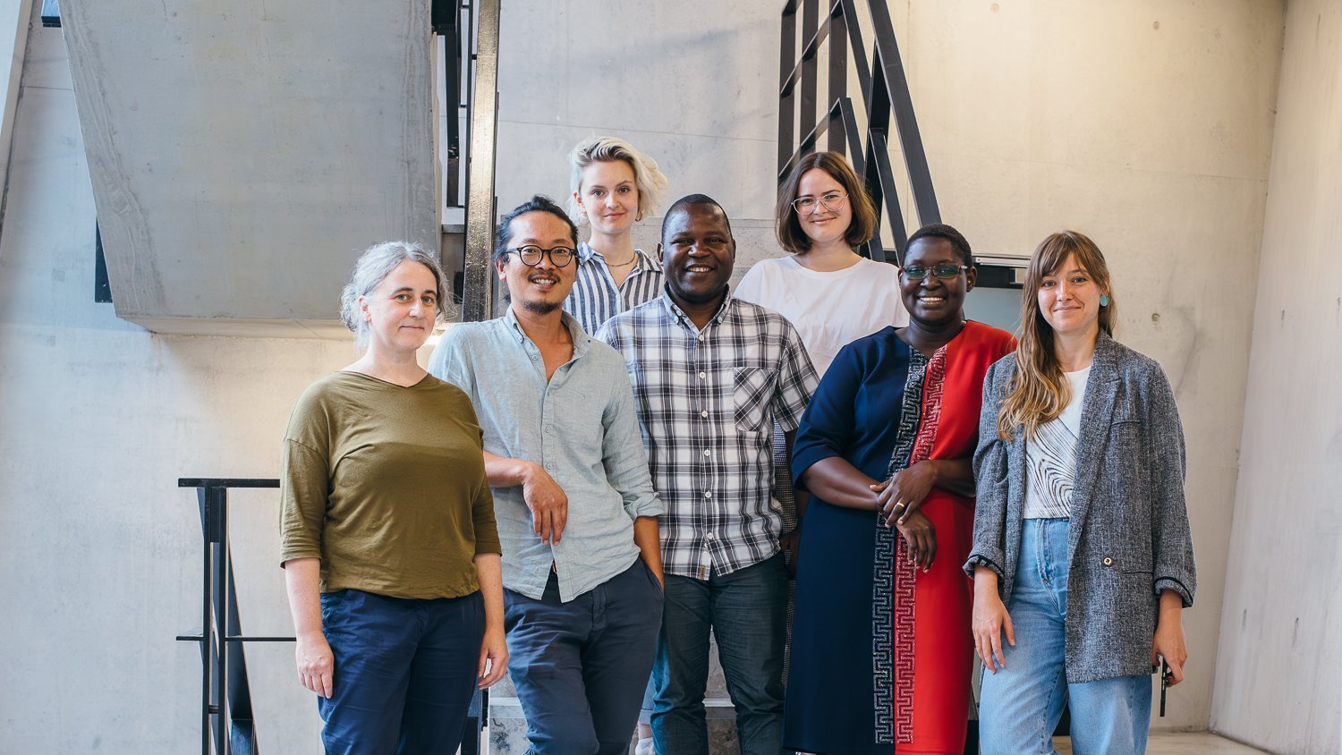 A group photo picturing seven diverse people standing in front of a staircase.
