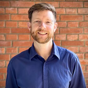 Dr. Benno Kreuels: a doctor with short reddish hair and a short reddish beard in a dark blue casual shirt stands in front of a brick wall. Above his left eyebrow he wears a piercing ring.