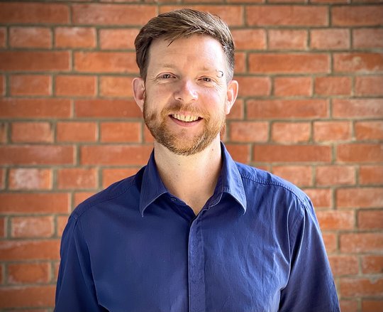 Benno Kreuels: a doctor with short reddish hair and a short reddish beard, wearing a dark blue casual shirt, stands in front of a brick wall. He wears a piercing ring above his left eyebrow.