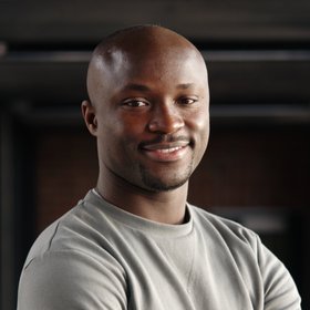 A project manager with a bald head, a short stubbly beard and a grey jumper. He has his arms folded in front of his chest and is standing in front of a dark background.