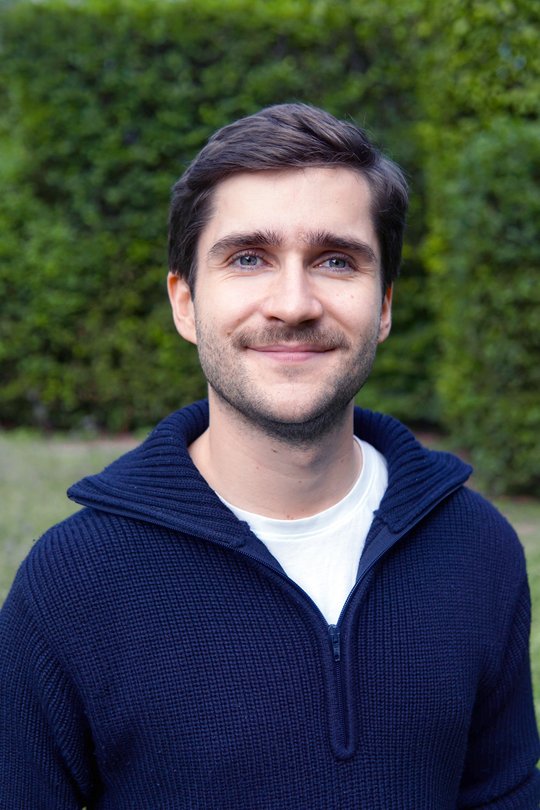 Kristopher Nolte Das Porträtfoto zeigt einen jungen Mann mit braunen Haaren, weißem T-Shirt und dunklem Pulli vor grünem Naturhintergrund.