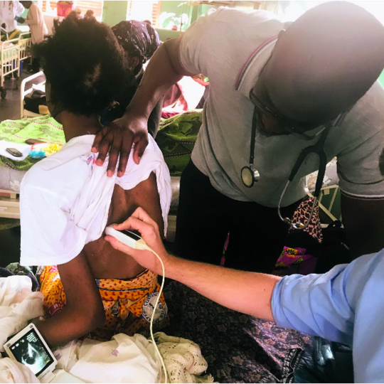 Point-of-Care Ultrasound (POCUS) in use in Malawi. The picture shows the treatment of a young patient in hospital. The patient is sitting on a bed with her back to the doctors. Two doctors are holding an ultrasound machine to her back while looking at a small monitor displaying the ultrasound image.