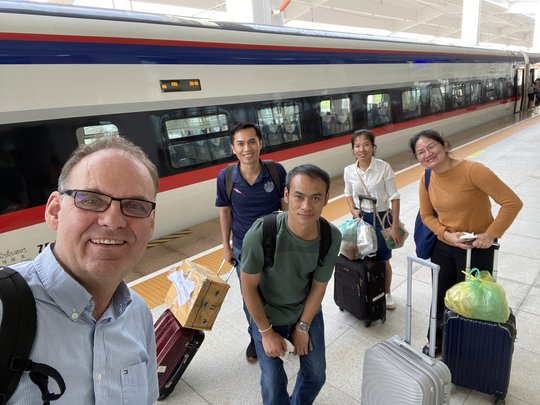 The picture shows five people standing in front of a train with luggage and looking friendly into the camera.