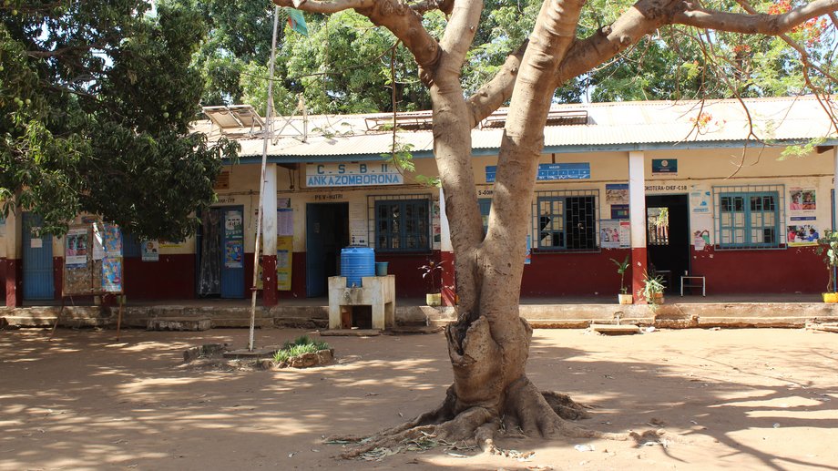 Gesundheitseinrichtung in Madagaskar Das Foto zeigt ein helles Gebäude mit grünen Bäumen im Hintergrund. Im Vordergrund ist ein großer brauner sich verzweigender Baumstamm zu sehen.