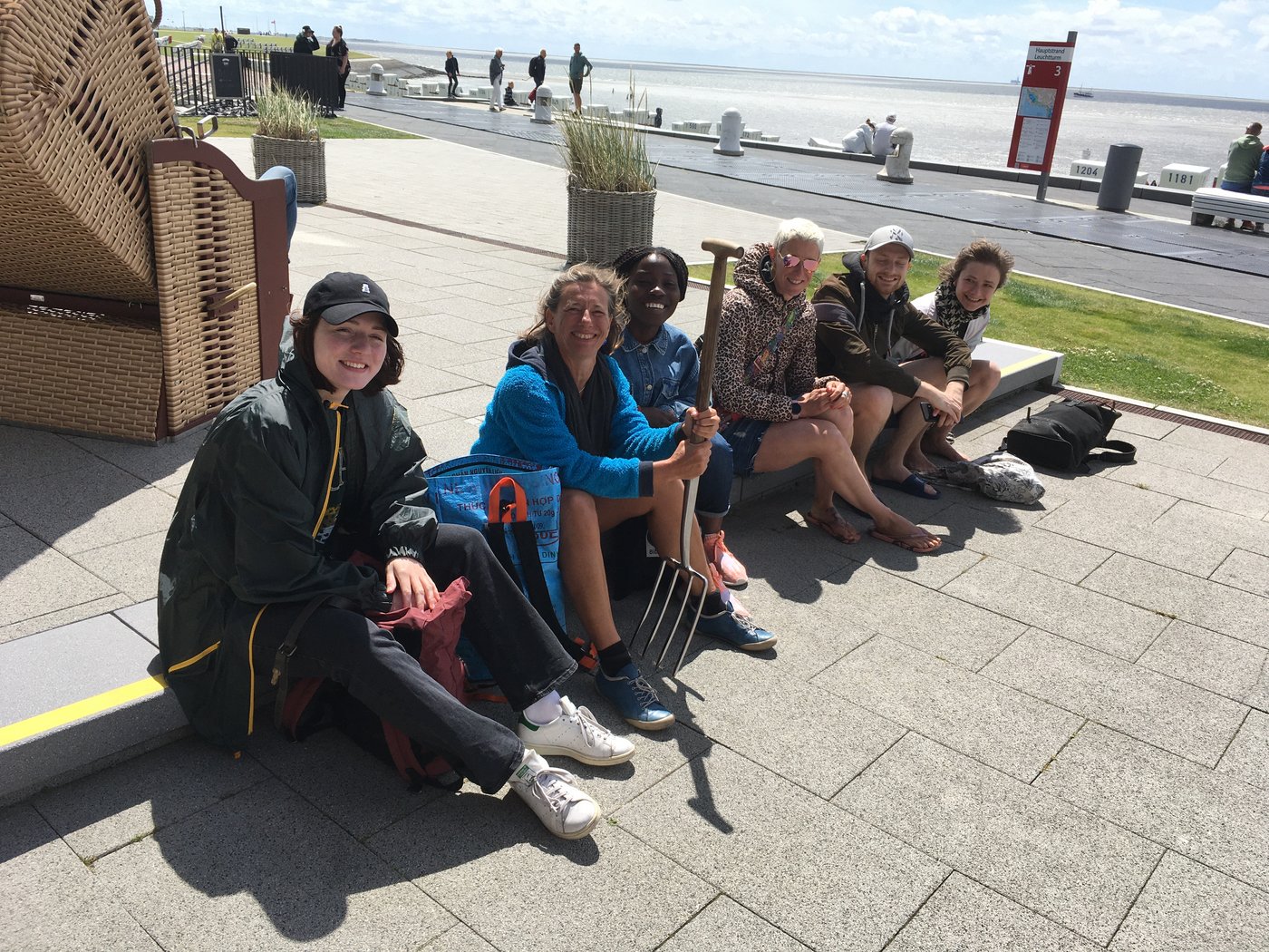 AG Helminthen-Immunologie an der Uferpromenade bei Büsum: Eine Gruppe von 6 Menschen sitzt an auf einem Kantstein und lachen ausgelassen in die Kamera. Eine Person hält eine Mistgabel in der Hand. Im Hintergrund kann man links einen Strandkorb sehen, dahinter die Uferpromenade und dahinter das Meer.