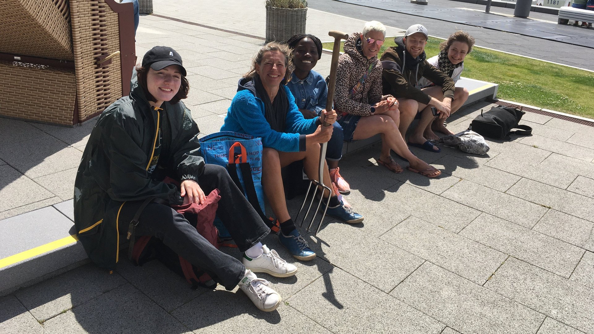 AG Helminthen-Immunologie an der Uferpromenade bei Büsum: Eine Gruppe von 6 Menschen sitzt an auf einem Kantstein und schauen in Richtung der Kamera. Eine Person hält eine Mistgabel in der Hand. Im Hintergrund steht links einen Strandkorb, dahinter die Uferpromenade und dahinter das Meer.