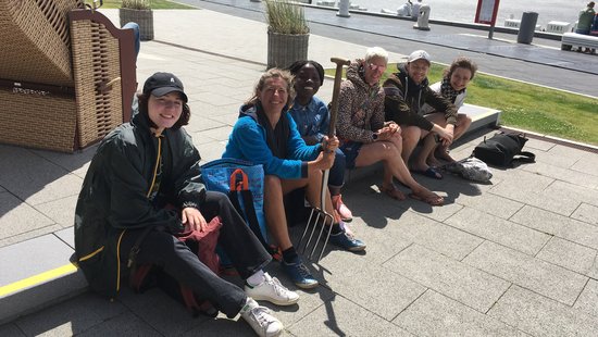 AG Helminthen-Immunologie an der Uferpromenade bei Büsum. AG Helminthen-Immunologie an der Uferpromenade bei Büsum: Eine Gruppe von 6 Menschen sitzt an auf einem Kantstein und schauen in Richtung der Kamera. Eine Person hält eine Mistgabel in der Hand. Im Hintergrund steht links einen Strandkorb, dahinter die Uferpromenade und dahinter das Meer.